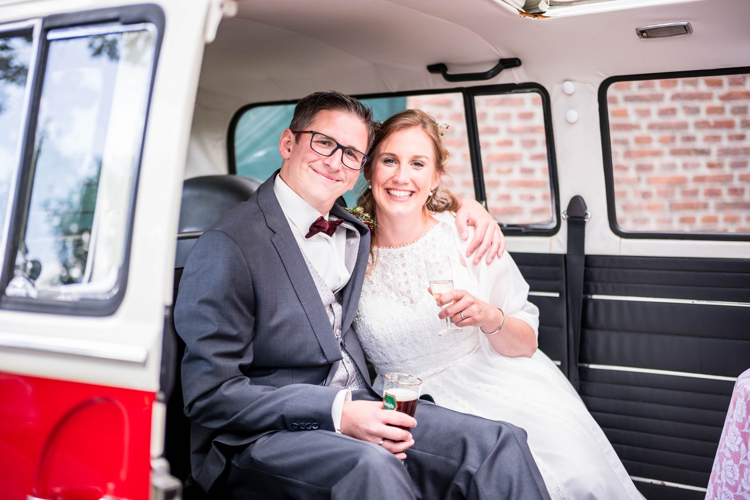 Ein fröhliches Brautpaar, das nach der Trauung Arm in Arm in einem alten, weiß-roten VW Bulli T2 sitzt und ein Sekt bzw. Alt Bier trinkt.