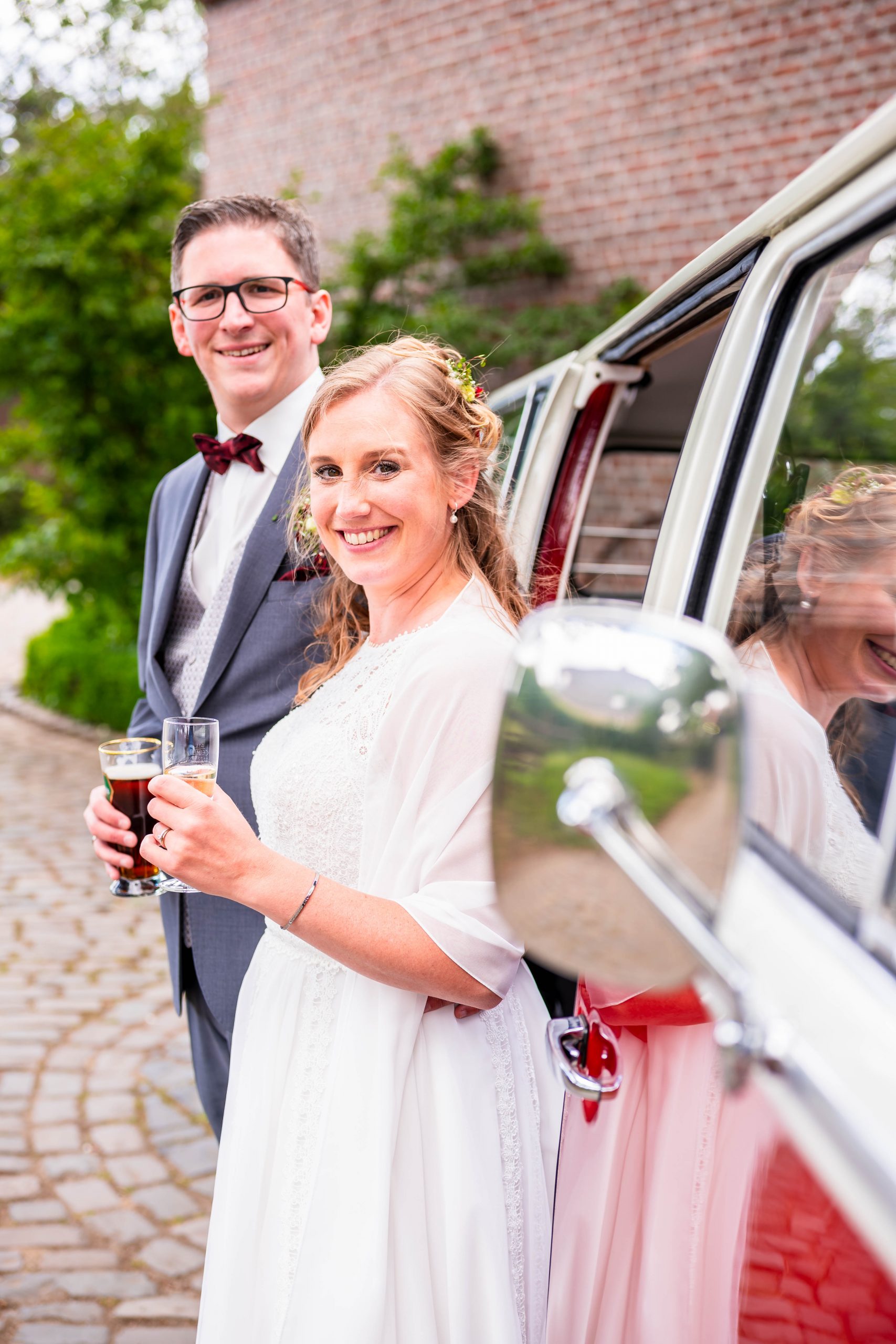 Ein fröhliches Brautpaar, dass neben einem weiß-roten alten VW Bulli T2 Bus mit geöffneter Türe steht.