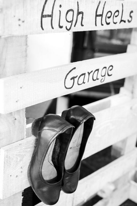 Schwarz-weiß Bild einer aufrecht stehenden Euro-Palette mit der Aufschrift "High Heels Garage" und einem schwarzen paar eingehängter Pumps, auf einer Hochzeitsfeier