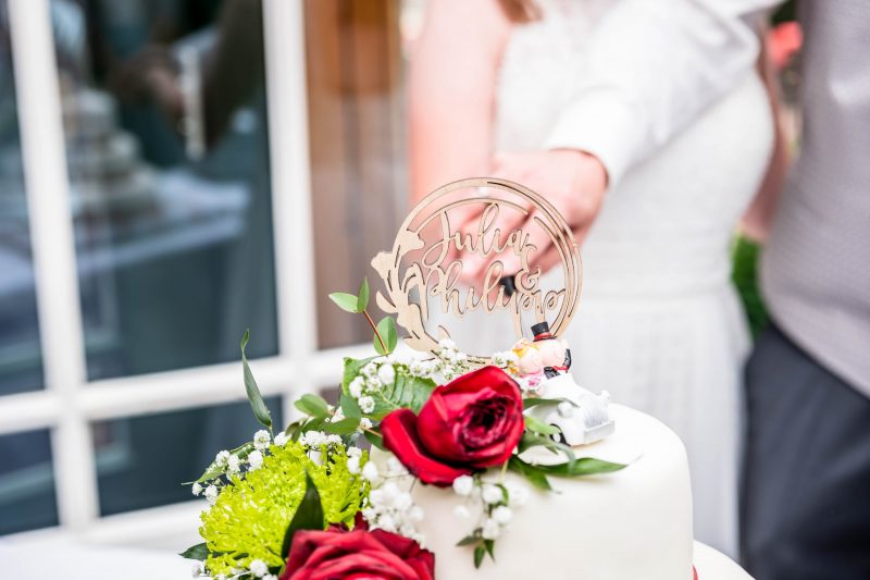 Eine weiße, mehrstöckige Hochzeitstorte mit roten Rosen und grünen Blättern wird vom Brautpaar gemeinsam angeschnitten.