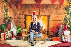 Ein kleiner Junge sitzt auf einem Schlitten vor einer Fotowand, die einen weihnachtlich geschmückten Kamin zeigt. Links und rechts sind Weihnachtswichtel und Geschenke.