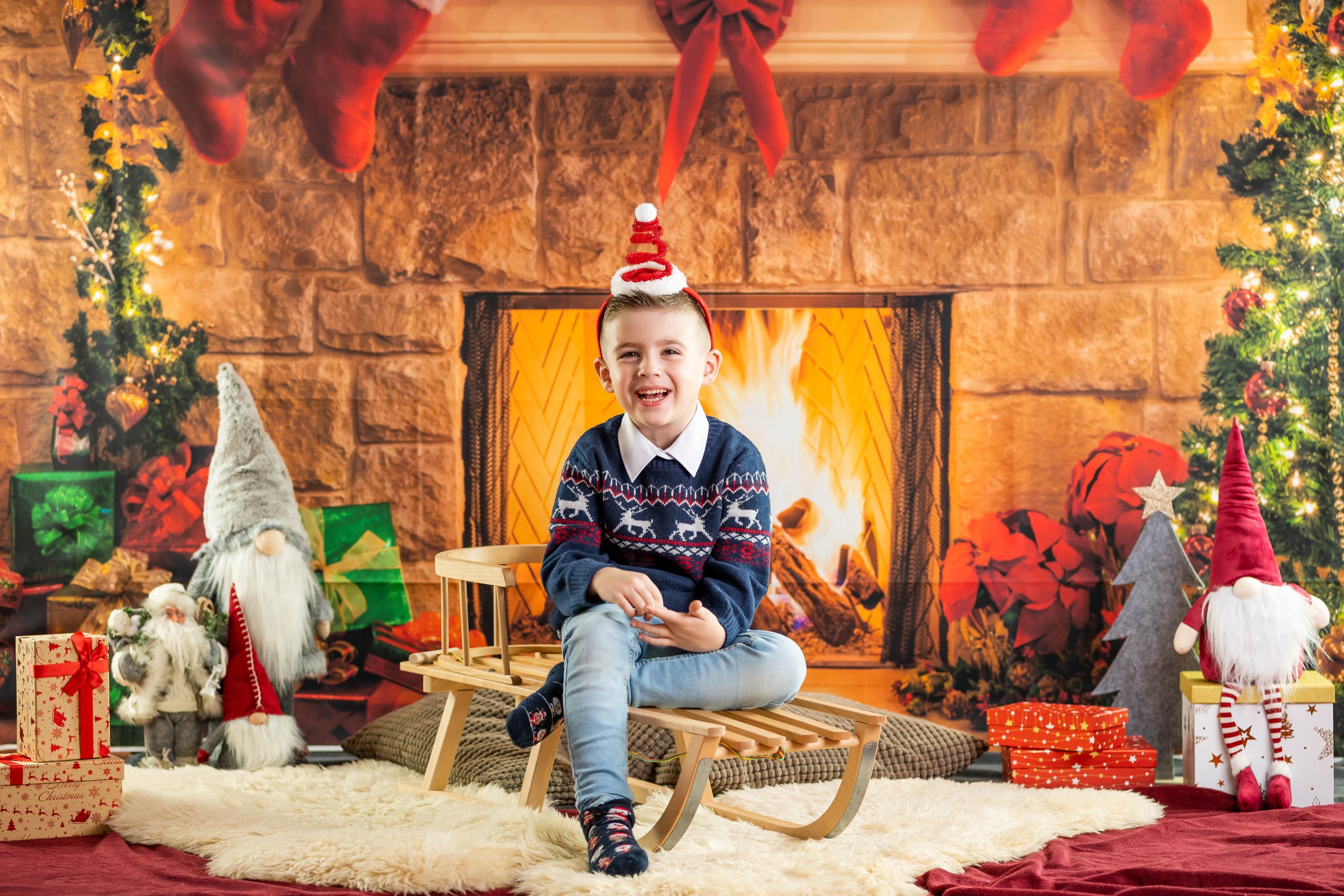 Ein kleiner Junge sitzt auf einem Schlitten vor einer Fotowand, die einen weihnachtlich geschmückten Kamin zeigt. Links und rechts sind Weihnachtswichtel und Geschenke.