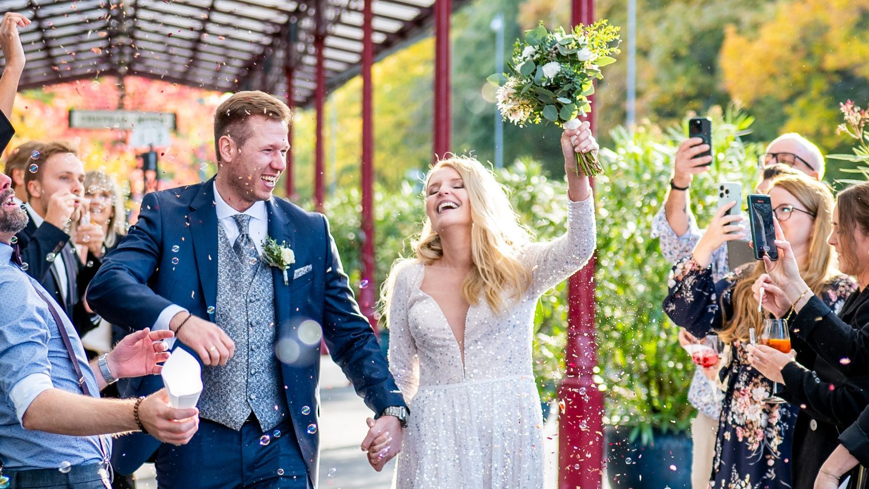 Ein Brautpaar erreicht den Empfang der Hochzeitsfeier im Nordbahnhof Krefeld. Die Braut hält jubelnd den Brautstrauß hoch, während die Gäste Blüten werfen und Seifenblasen pusten.