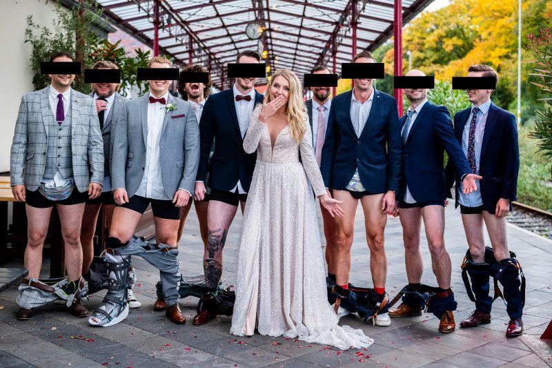 Eine schockiert guckende Braut, die in der Mitte zwischen männlichen Hochzeitsgästen steht, die alle ihre Hose heruntergelassen haben. Witziges Hochzeitsfoto. Auf dem Bahnsteig des Nordbahnhof Krefeld.